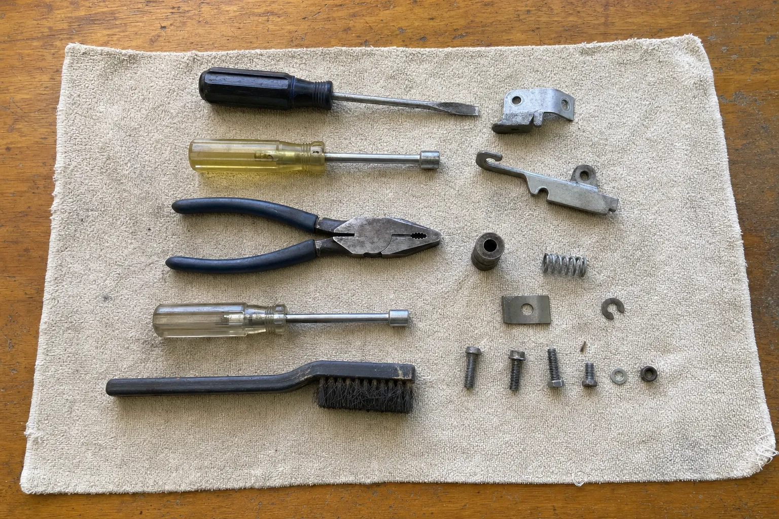 A hooked lever, a formed bracket, a coil spring, a bushing and a clip beside a flat screwdriver, two nut drivers, pliers and a wire brush