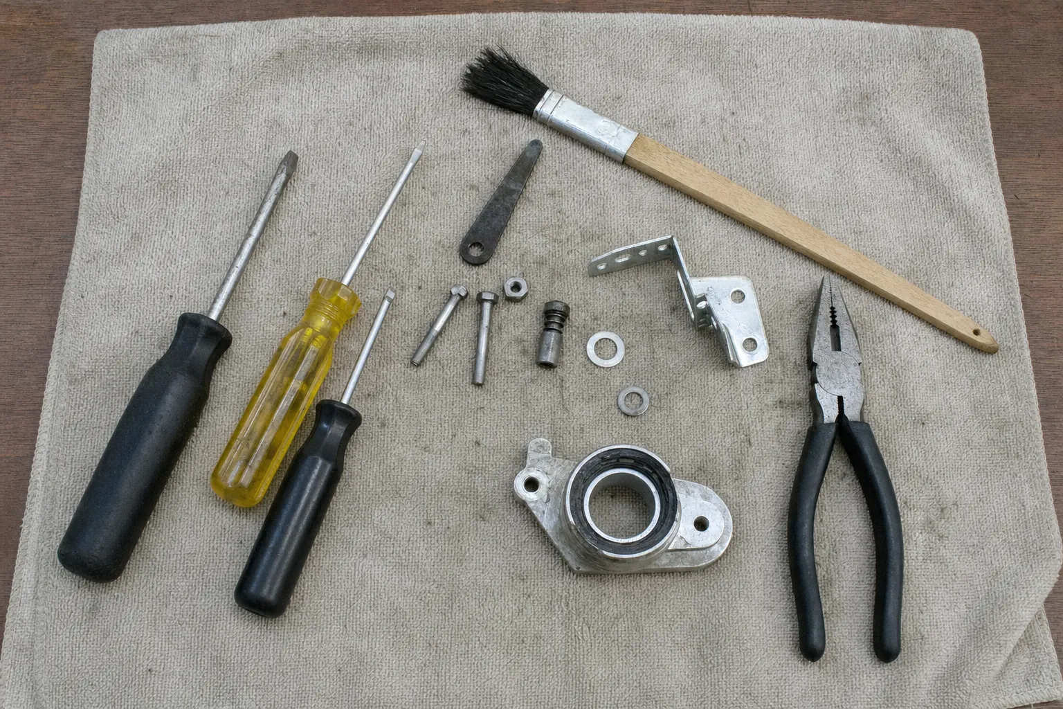 A flanged bearing housing with its seal, a bent bracket, bolts and washers laid out with three screwdrivers, pliers and a brush