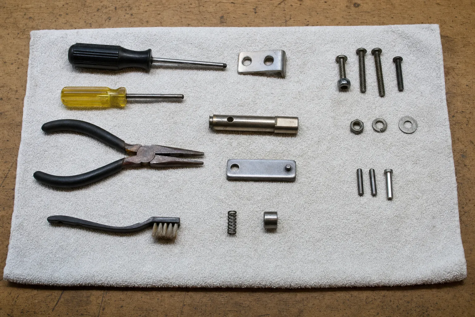 A short drilled shaft, a flat link plate, a small spring, a bushing and pins set out in rows beside screwdrivers and needle-nose pliers